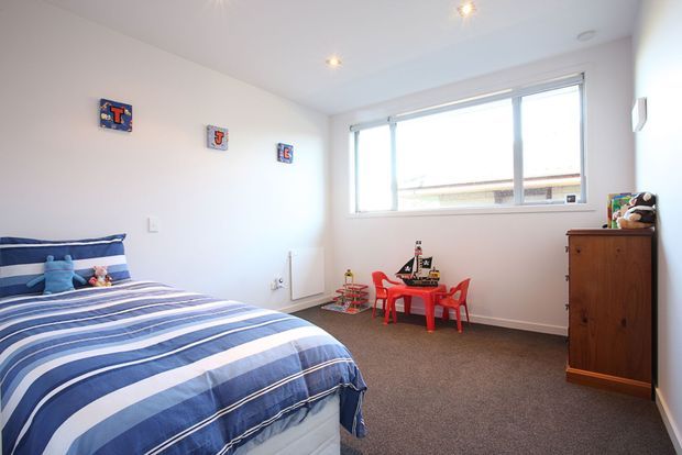 Bright children's bedroom with a blue-and-white striped bed, colorful wall decorations, a small red children's table and chairs with toys, a wooden dresser, white walls, and carpeted flooring. The room has large horizontal windows and recessed ceiling lights.
