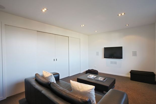 A clean, contemporary media or living room with a large black leather sectional sofa, a low black rectangular coffee table, wall-mounted television, concealed white built-in cabinetry, recessed downlights, and neutral carpeted flooring. The room has a minimalist, uncluttered appearance with white walls and integrated speakers.