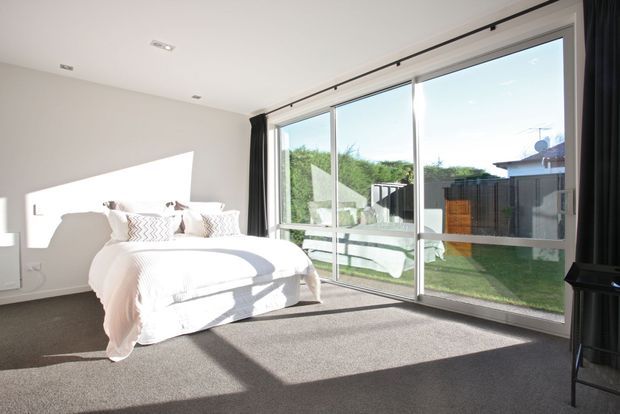 Bright contemporary bedroom with a large white bed, soft white bedding, dark carpet, and full-height sliding glass doors opening to a small garden courtyard. Strong sunlight creates geometric shadows across the walls and floor, while dark curtains frame the glazing.