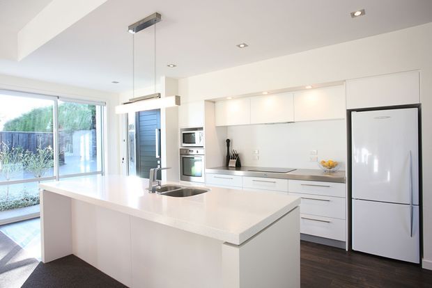 Bright contemporary white kitchen with a large island featuring an integrated sink and waterfall-style end panel. The room has sleek white handleless cabinetry, a pale benchtop, stainless-steel appliances, dark timber flooring, recessed downlights, and large glazed doors opening to an outdoor area.