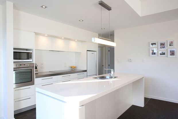 Bright contemporary kitchen with white flat-panel cabinetry, integrated stainless-steel appliances, a large white island with sink and tap, dark benchtop accents, recessed ceiling lights, and a linear pendant over the island. The kitchen opens toward a white-walled living or dining area with framed photographs and dark carpet flooring.