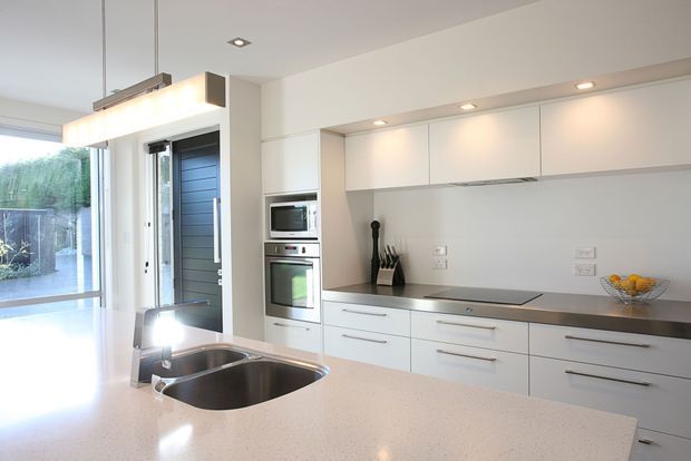 Bright contemporary kitchen with a large light-coloured island, integrated double sink, flat-panel white cabinetry, pale stone benchtops, stainless-steel appliances, and a dark induction cooktop. Recessed ceiling lighting and a linear pendant illuminate the space, while full-height glazing and a glazed door connect to the exterior.