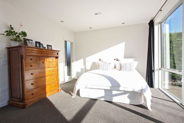Bright contemporary bedroom with a white upholstered bed, patterned throw pillows, dark carpet, and a traditional dark timber chest of drawers with framed photographs and a potted plant. Large floor-to-ceiling glazed doors and windows with black curtains bring in strong sunlight and overlook greenery.