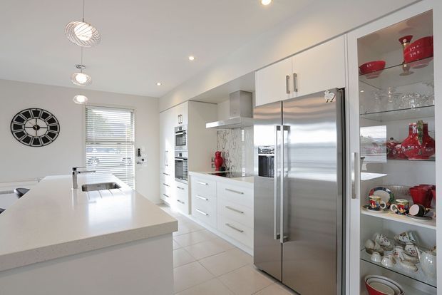 Bright, clean contemporary kitchen with a large white island, integrated white cabinetry, stainless-steel double-door refrigerator, built-in ovens, glass-front display cabinetry, and tiled flooring. A dining area and window with blinds are visible beyond the island.