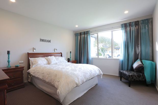 Bright, comfortably furnished bedroom with a wooden bed and matching bedside furniture, white textured bedding, patterned carpet, and a large window dressed with blue-teal curtains. A pair of upholstered occasional chairs sits near the window, and recessed ceiling lights provide additional illumination.