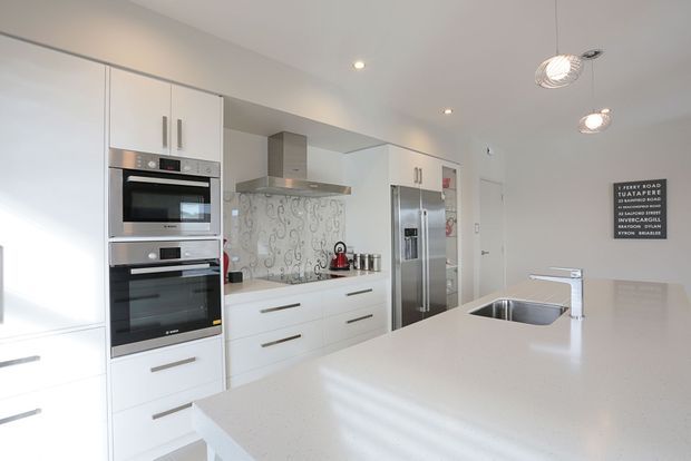 Bright contemporary white kitchen with extensive built-in cabinetry, integrated stainless-steel appliances, a large pale engineered-stone island with undermount sink, and a decorative patterned splashback. Recessed ceiling lights and pendant fixtures provide a clean, refined finish.