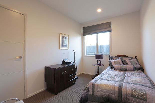 Compact, softly lit bedroom with a single bed, patterned quilt and pillows, dark timber dresser, framed artwork, carpeted floor, and a window with a roller blind. The room has neutral painted walls and simple contemporary-traditional furnishings.