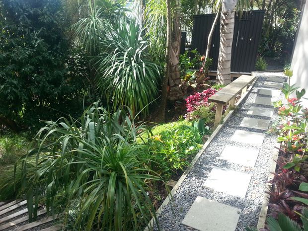 A lush tropical garden walkway with large rectangular stepping stones set into a grey gravel path. Dense foliage, palms, colourful ornamental plants, a simple timber bench, and dark vertical screening create a secluded, resort-like outdoor passage.