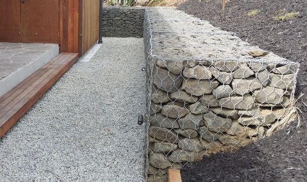 Close-up exterior view of a landscaped building entrance, showing a gravel walkway beside timber-clad walls and a raised gabion retaining wall filled with irregular natural stone. Timber steps and a partially visible wooden door are at left, with dark mulch and planting above the retaining wall.