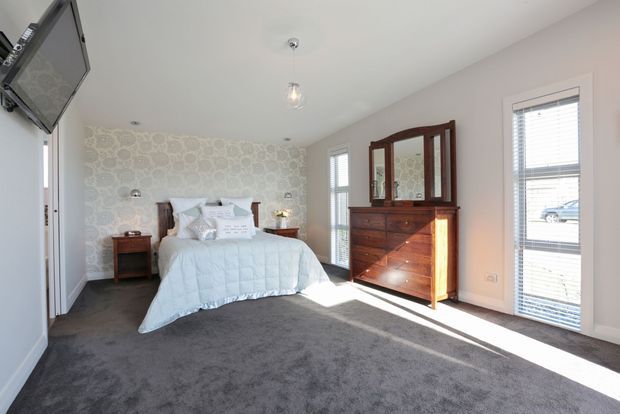Bright bedroom with a centered bed, dark grey carpet, pale walls, patterned feature wallpaper behind the bed, and tall windows with white blinds. A traditional dark timber dresser with mirror and matching bedside tables provide warm contrast, while a wall-mounted television and simple pendant light complete the room.