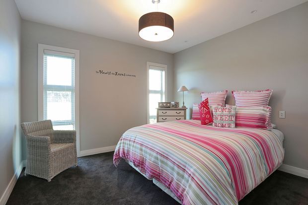 Bright, comfortably furnished bedroom with two tall windows, pale grey walls, dark carpet, a wicker armchair, a small dresser, and a bed dressed in colourful pink, white, and grey striped bedding with decorative pillows. A drum pendant light hangs from the ceiling, and a small wall inscription adds a playful personal touch.