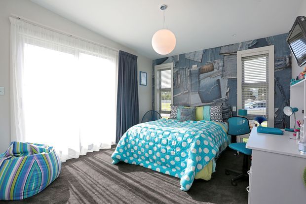 A bright, playful bedroom with a double bed dressed in a turquoise-and-white polka-dot duvet, a blue striped beanbag, dark grey carpet, sheer curtains, and navy drapes. The feature wall has blue-grey patchwork-style wallpaper, with two shuttered windows, a turquoise desk chair, a white desk and storage cabinetry, and a round pendant light.