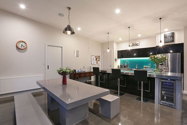 Open-plan contemporary kitchen and dining area with a large pale stone island and integrated bench seating in the foreground. A black kitchen island with bar stools, glossy dark cabinetry, stainless-steel appliances, and a turquoise illuminated splashback occupies the rear. Pendant lights, recessed ceiling lights, polished flooring, and minimal white walls create a refined entertaining space.