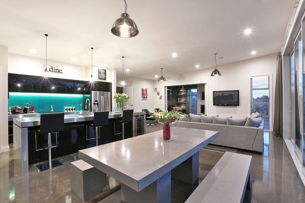 Open-plan contemporary kitchen, dining and living area with a black kitchen island, turquoise glass splashback, stainless-steel appliances, polished concrete-style flooring, a large stone-look dining table with bench seating, grey sectional sofa, pendant lights and large windows.