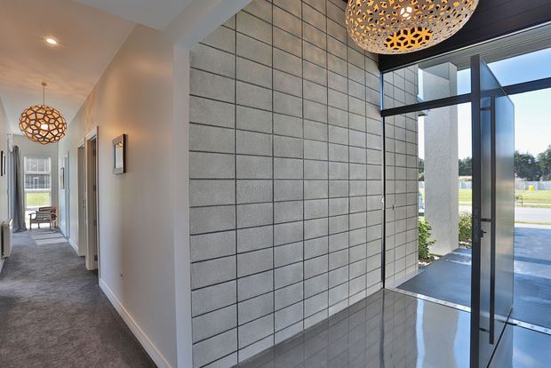 A bright contemporary entryway and hallway featuring a large grey tiled feature wall, polished reflective floor tiles, white walls, charcoal-framed floor-to-ceiling glazing, and a modern geometric pendant light. The hallway extends toward a furnished room at the rear, while the glazed entrance opens to the driveway outside.