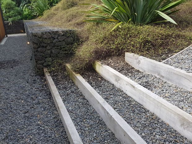 A landscaped exterior walkway with stepped gravel paths and pale timber or concrete retaining sleepers, bordered by dark stone gabion-style retaining walls and dense ornamental planting. The design has a natural, structured garden character with subtle pathway lighting.