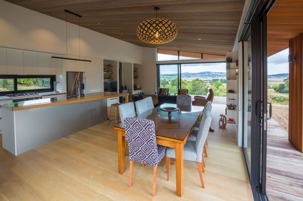 A bright open-plan kitchen and dining area with light timber flooring and a warm timber ceiling. The kitchen has sleek white full-height cabinetry, a long island with a timber benchtop, stainless-steel appliances, and a dark horizontal splashback window. A large timber dining table with upholstered chairs occupies the centre, beneath a decorative woven pendant light. Full-height sliding glass doors open to a timber deck and a broad rural or semi-rural landscape beyond.