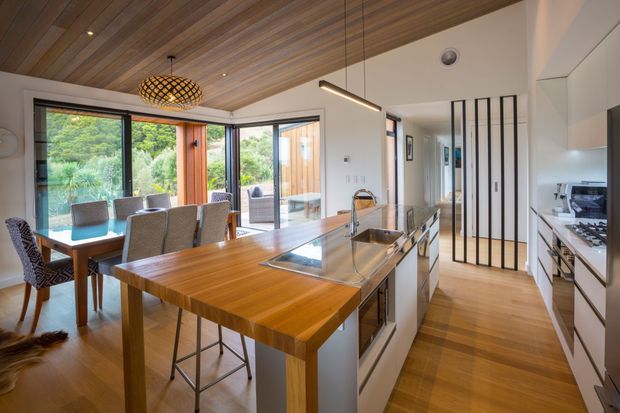 A bright, open-plan kitchen and dining area with extensive timber detailing. The kitchen features a large timber island with integrated cooktop and sink, light cabinetry, and a stainless-steel benchtop. A timber dining table sits beside floor-to-ceiling glazing and opens toward a leafy outdoor area. The ceiling is clad in warm timber boards, complemented by timber flooring, black-framed glazing, and a vertical black metal screen near the hallway. Contemporary, warm, and natural in character.