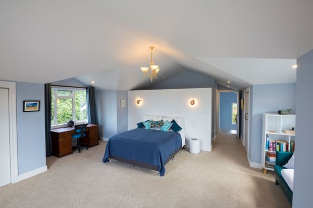 A spacious attic-style bedroom with a central bed, pale blue-grey walls, beige carpet, and a sloped ceiling with a decorative chandelier. The bed is positioned against a partial white partition wall with small wall lights, while a desk and timber drawer units sit beside a window overlooking greenery. An open doorway leads to a hall or adjoining space, and a white shelving unit provides storage on the right.