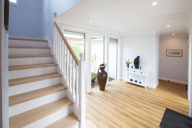 Bright residential entry hall with a timber staircase, white-painted balustrade and walls, light timber flooring, tall glazed doors and windows, and a small white console displaying decorative objects. The stair rises beside an open landing with a high, airy ceiling and recessed downlights.