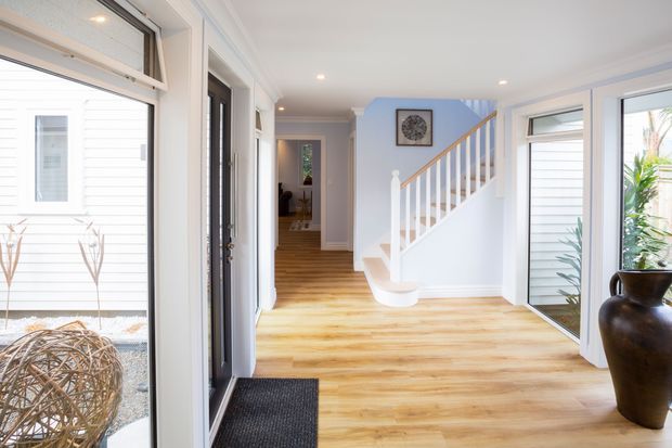 Bright residential entry hall with pale timber flooring, white walls and trim, large glazed openings, and a white staircase with timber handrail and white balusters. A dark entry door and mat provide contrast, while a corridor leads toward rooms at the rear. The space has a clean, airy, contemporary coastal character with abundant natural light.