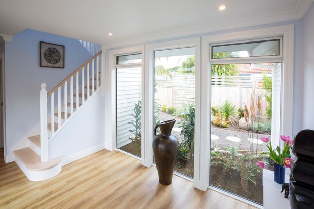 Bright residential stair landing or entry area with a white-painted staircase, pale blue walls, light timber-look flooring, and a bank of large white-framed windows overlooking a landscaped garden. A tall decorative vase and indoor plant add minimal decor, while recessed ceiling lights provide even illumination.