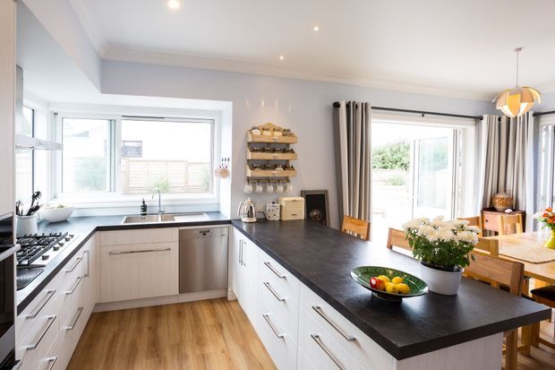 Bright open-plan kitchen and dining area with white shaker-style cabinetry, a dark stone-look island benchtop, timber-look flooring, stainless-steel appliances, and large windows and glazed doors providing abundant natural light. A timber dining table and chairs are visible beyond the kitchen, with open shelving and neutral curtains adding warmth.