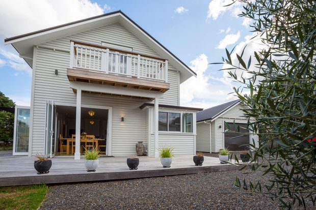 Exterior view of a light-coloured two-storey weatherboard house with a pitched gable roof, upper balcony, covered ground-floor porch, extensive glazing, and a broad timber deck. A gravel forecourt, potted plants, and a neighbouring matching structure are visible; the image has a bright, relaxed residential character.
