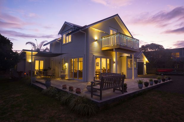 Twilight exterior view of a multi-level coastal-style home with grey horizontal weatherboard cladding, steep gabled roofs, extensive warm-lit glazing, and a raised timber deck. The deck includes a small upper balcony with white railings, outdoor seating, potted plants, and landscaped grasses around the house.