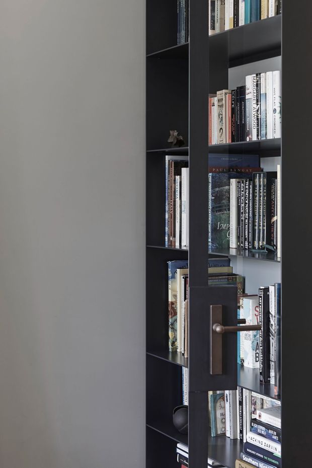 Close-up interior view of a tall, dark built-in bookcase filled with books. A narrow hinged or pivoting shelving door is partially closed in front of the bookcase, featuring a dark rectangular panel and a horizontal metal lever handle. The surrounding wall is smooth grey, creating a restrained, contemporary library-like composition.