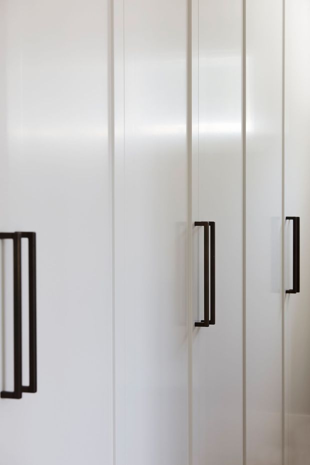 Close-up of tall white built-in cabinetry with clean, flat-panel doors and slim dark rectangular bar handles. The minimalist joinery fills the frame, with soft natural lighting and little surrounding context.