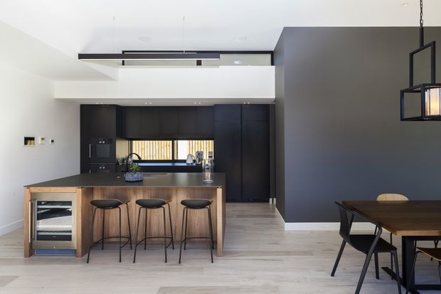 A contemporary open-plan kitchen and dining area with a large timber island, integrated dark cabinetry, black pendant lighting, pale timber flooring, and a dark charcoal feature wall. Three bar stools line the island, while a dining table is partially visible in the foreground.