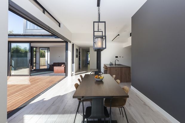 Bright contemporary open-plan dining and kitchen area with a long dark timber dining table, pendant light, compact timber kitchen cabinetry, and large black-framed sliding glass doors opening to a timber deck and adjoining living area. The space features a high ceiling, abundant natural light, and a strong indoor-outdoor connection.