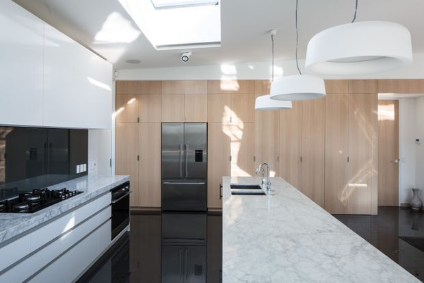 Bright contemporary kitchen with a long white marble island, integrated stainless-steel sink and tap, white handleless cabinetry, black glass splashback, black appliances, and a full-height light timber storage wall. Large pendant lights and a skylight provide strong natural illumination, while dark flooring creates contrast.