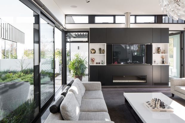 Bright contemporary living room with floor-to-ceiling black-framed glazing opening toward a landscaped outdoor area. A large dark built-in media wall incorporates a television, open display niches, and a linear fireplace. Pale sectional and armchairs surround a white coffee table with a chess set, while a sculptural pendant light adds a focal accent.