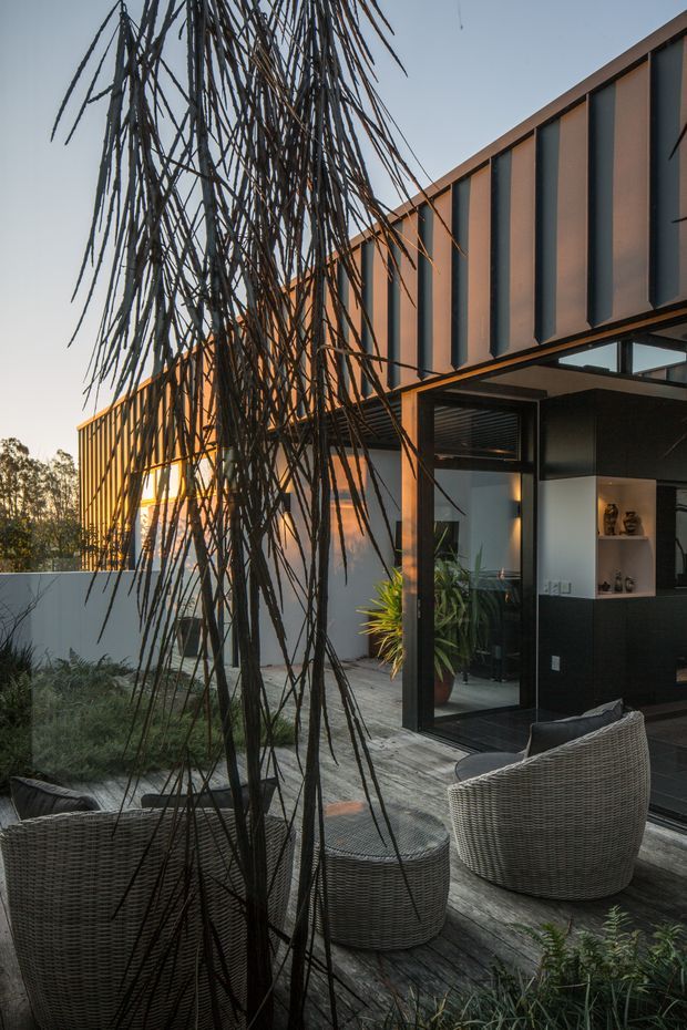 A contemporary residential exterior viewed from a landscaped outdoor sitting area. The house features a long, angular form with dark vertical metal cladding panels, warm timber-toned framing, extensive black-framed glazing and an open corner connection to the interior. A weathered timber deck, woven outdoor armchairs and a low table form the foreground, with structured planting and a sculptural slender tree creating a natural screen.