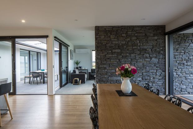 Open-plan dining area with a long timber dining table and black sculptural chairs, centred against a prominent dark stacked-stone feature wall. Large dark-framed glazed sliding doors connect the room to a bright courtyard or sunroom, while a living area is visible beyond. Pale timber flooring, white walls and recessed ceiling lights create a contemporary, open and light-filled interior.