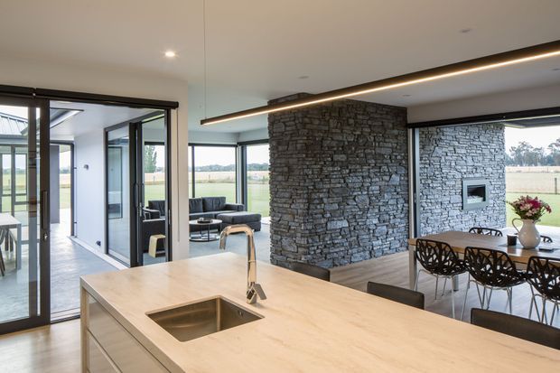 A bright, open-plan kitchen and dining area with a large pale timber island, integrated rectangular sink and brushed metal tap. Full-height black-framed glazing and sliding doors open to a rural landscape. A dark stacked-stone feature wall with a built-in fireplace divides the kitchen from the dining and lounge areas. Linear recessed lighting, neutral finishes, and contemporary furniture create a refined modern interior.