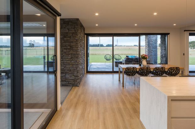 Open-plan contemporary kitchen, dining, and living area with extensive floor-to-ceiling glazing overlooking flat rural farmland. The space features a large pale stone kitchen island, light timber flooring, dark-framed sliding glass doors, exposed stacked-stone feature piers, recessed ceiling lighting, and a dining table with sculptural black chairs. An outdoor seating area is visible beyond the glazing.