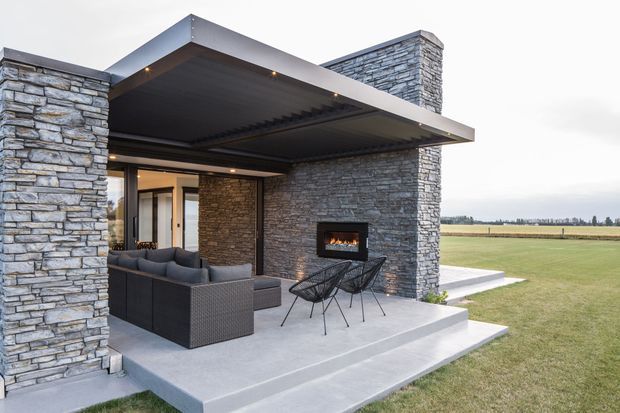 Contemporary single-storey outdoor entertaining area with a large flat-roofed cantilevered canopy, grey stacked-stone walls and chimney, concrete terrace, built-in fireplace, outdoor lounge seating, and open views across a flat rural landscape.