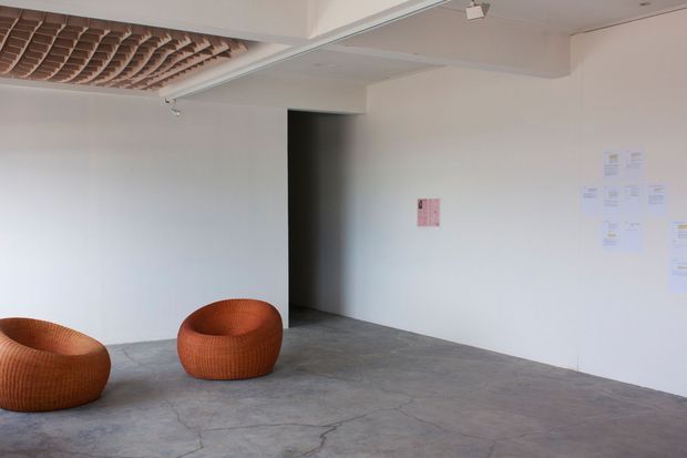 A sparse, gallery-like interior with white walls, a polished grey concrete floor, and two low rounded woven lounge chairs in warm terracotta tones. A dark open doorway punctuates the rear wall, while a suspended sculptural timber slat ceiling feature occupies the upper left. Small papers are mounted on the wall, suggesting an exhibition or commercial display space.