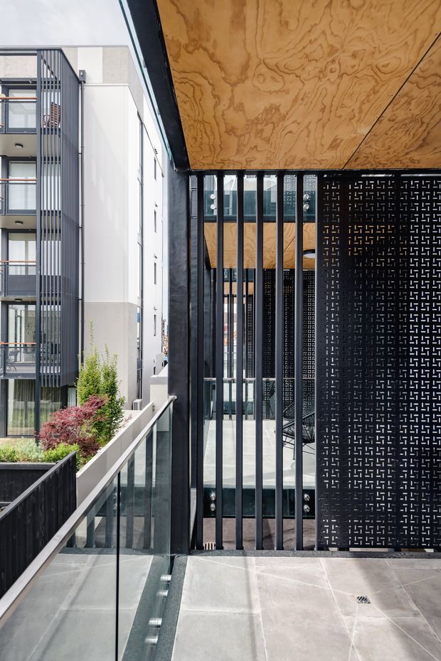 A contemporary multi-storey residential or mixed-use building viewed from an elevated external walkway or balcony. The foreground features a covered passage with a plywood-lined soffit, black steel framing, vertical metal security screens, perforated black privacy panels, and glass balustrades. Adjacent white and grey building facades with glazed balconies are visible, along with integrated planter beds and low landscaping. The composition highlights layered circulation spaces, screens, and facade detailing in a bright urban setting.