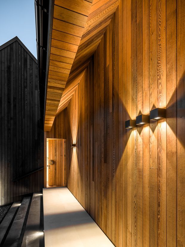 A narrow, contemporary exterior walkway between two dark-clad building volumes. The main facade is richly detailed with vertical timber wall cladding and a projecting timber-lined soffit, illuminated by black modern wall sconces. Dark steps and black vertical cladding contrast with the warm timber facade and pale paved walkway.