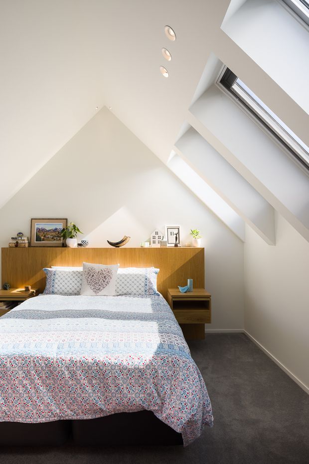 Bright attic-style bedroom with a pitched ceiling and multiple skylights, creating strong natural illumination. The room features a neatly made bed with patterned blue, white and red bedding, a warm timber headboard and integrated bedside storage, framed artwork and small decorative objects. Dark grey carpet covers the floor, while the walls and ceiling are painted white.