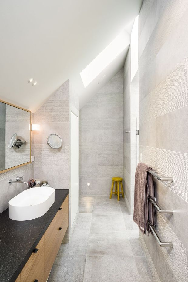 Bright contemporary bathroom with a walk-in shower, pale textured wall and floor tiles, a floating timber vanity with a white vessel basin, dark stone-look benchtop, large mirror, skylight, and a chrome heated towel rail. A small yellow stool adds a vivid accent.