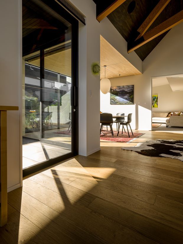 Sunlit interior view from an entry or circulation space into an open-plan dining and living area. The room features warm timber flooring, a dramatic exposed pitched timber ceiling with structural beams, large black-framed glazed doors opening to a planted courtyard, a dining table with dark chairs, and a partially visible lounge. Strong afternoon shadows create a warm, inviting atmosphere.