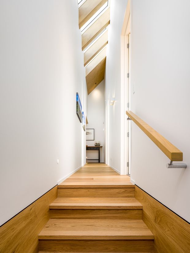 Bright, narrow residential stairway and hallway with warm timber stairs, timber wall panelling along the lower walls, white walls, and a matching timber handrail on a metal bracket. The stairwell rises to a long corridor beneath a sloping timber-lined ceiling with narrow skylights, creating an airy vertical connection. Minimal artwork and a small console are visible at the far end.