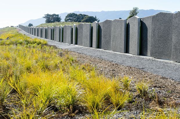 Exterior landscape view of a long, modern grey textured concrete privacy wall with repeated projecting vertical panels, set beside a gravel path and native ornamental grasses. Mountains and mature trees form the distant background.