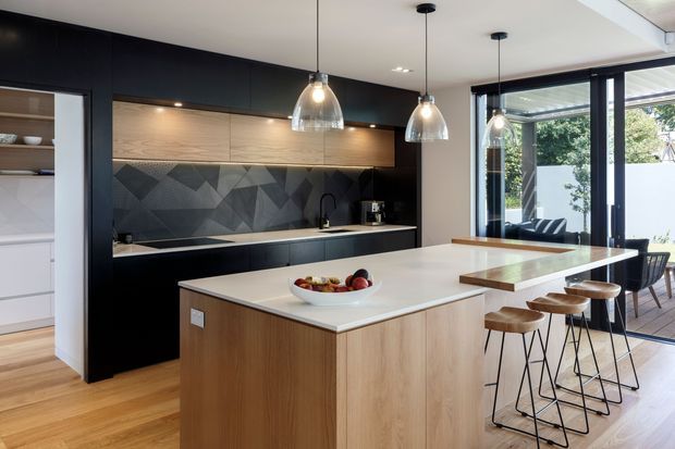 A spacious contemporary kitchen with a large timber island and white benchtop, matte black cabinetry, a warm timber overhead feature, and a geometric dark splashback. Three glass pendant lights hang above the island, which includes casual bar seating. A dining area and glazed doors connect to an outdoor space, while light timber flooring runs throughout.