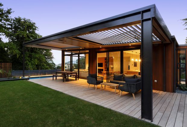 A modern single-storey home opening onto a generous timber deck and lawn. The outdoor entertaining area features a black steel-framed pergola with slatted timber soffit and louvred roof, built-in lighting, an outdoor dining table, and a lounge setting. Full-height glazing connects the deck to a warmly lit interior living and dining space, with a swimming pool visible beyond.